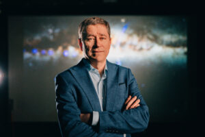 A picture of Matthew Bailes used as the hero image. Matthew is wearing a blue suit and has his arms crossed while smiling. Behind him is a projection of space and stars.