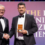 picture of two men standing side by side shaking hands. The man on the right is holding a medal.